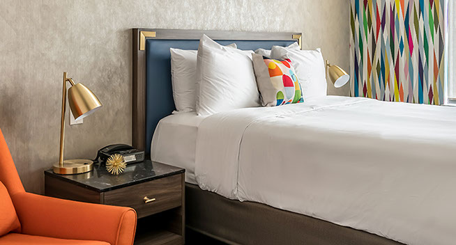 This photo shows the colorful ambience with a queen size bed and an orange sleeper chair, nightstand and lamp with alarm clock.