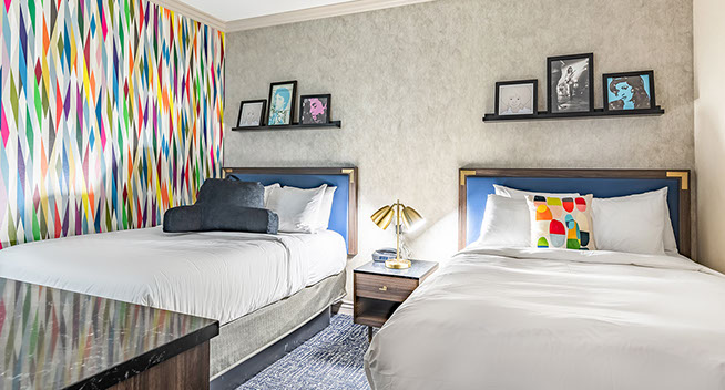 View of room showing to full size beds with  a trendy style decorated room with colorful wall paper and pillows. Night stand and lamp.
